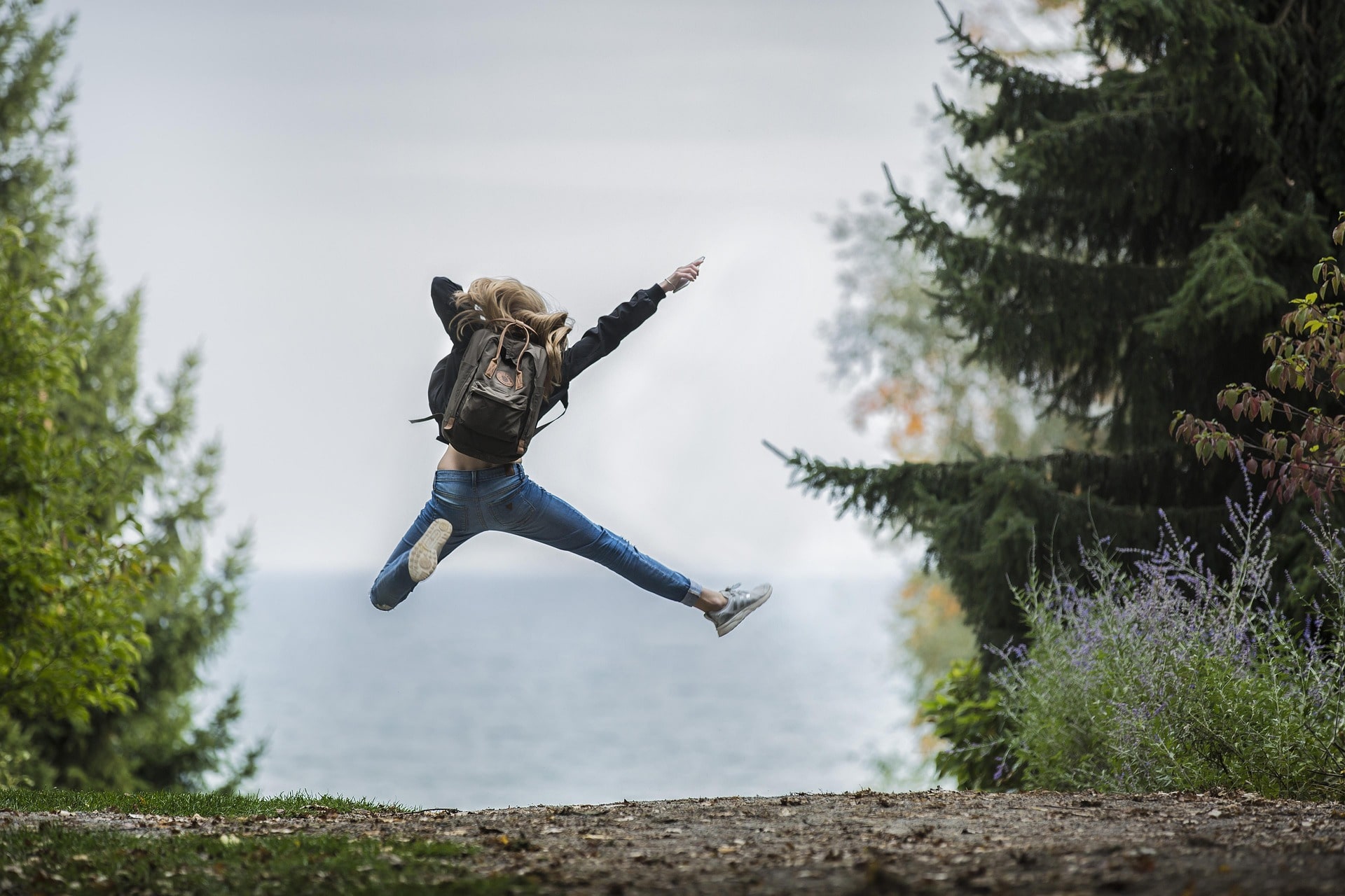 Foto einer jungen Frau, welche einen Luftsprung macht zwischen Bäumen und auf einen See blickt